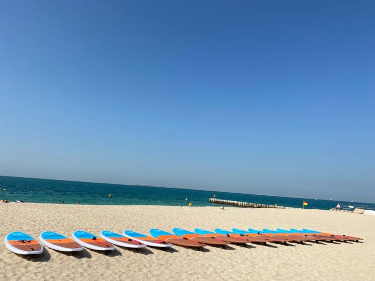 Water sports equipment bulk supply B2B: rows of paddle boards lined up on a sandy beach with a clear blue sky and calm ocean in the background in Dubai.