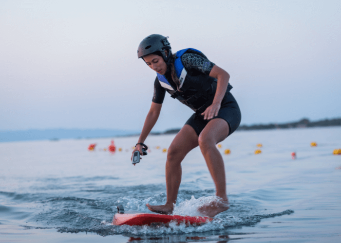 Person riding an electric foil board wearing a helmet and life jacket on calm waters.